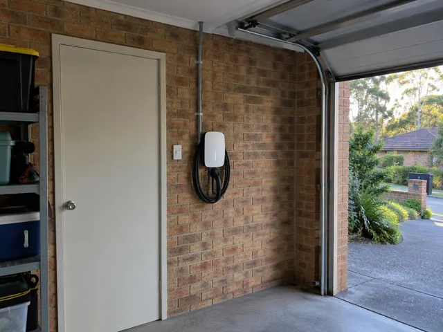 Wall-mounted EV charger on a brick garage wall beside a door and shelving in a home garage in Frenchs Forest